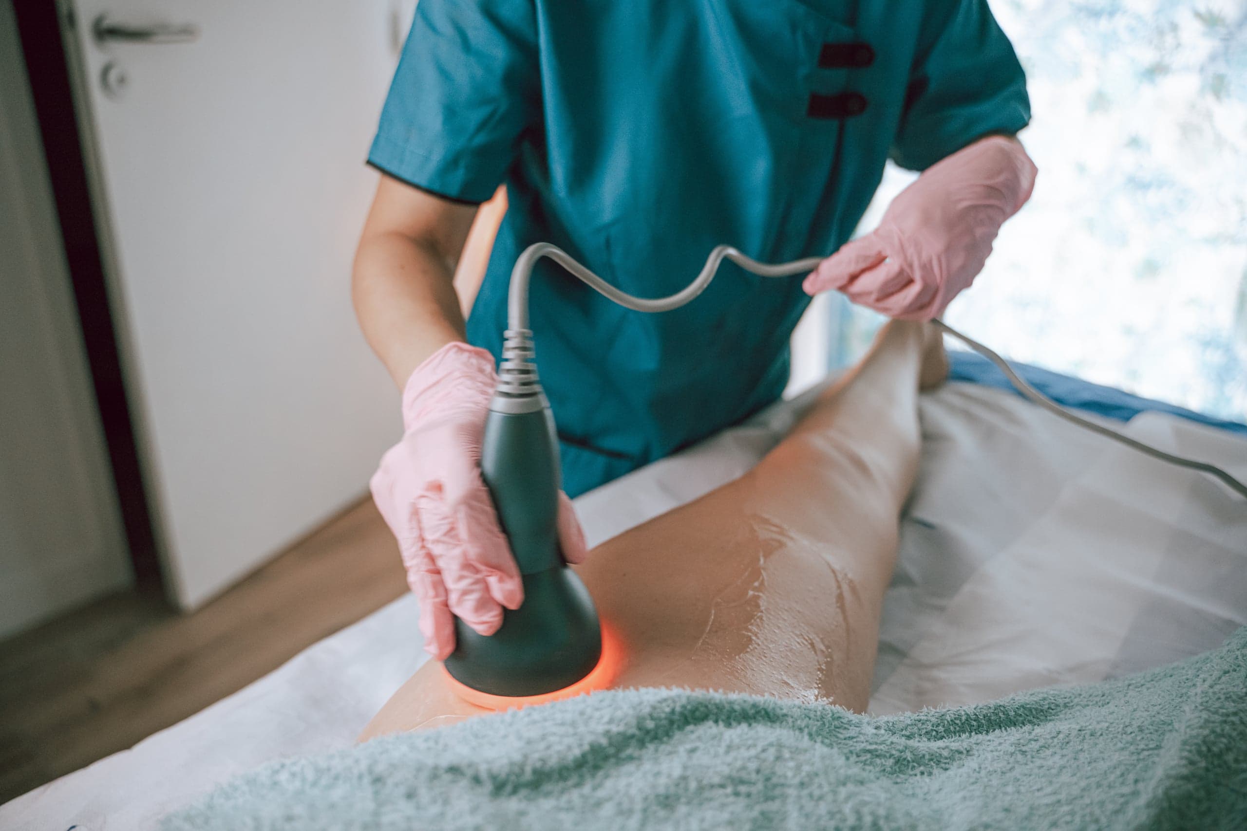 Tête de lipocavitation à lumière rouge appliquée sur la cuisse d'une cliente, gants de soin et tunique turquoise, cabinet de Vertou